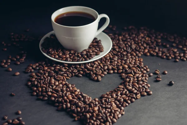 Taza con una bebida fragante sobre un fondo oscuro corazón de granos de café platillo blanco — Foto de Stock