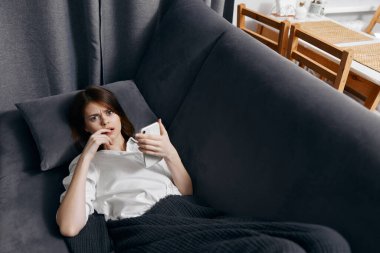 Kadın evin içinde kanepeye uzanmış, elinde cep telefonu var.