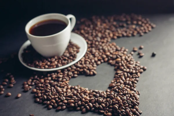 Taza con una bebida fragante sobre un fondo oscuro corazón de granos de café platillo blanco — Foto de Stock