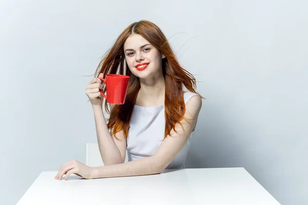 Attraktiv rödhårig kvinna dricker ur en röd mugg sittande vid bordet — Stockfoto