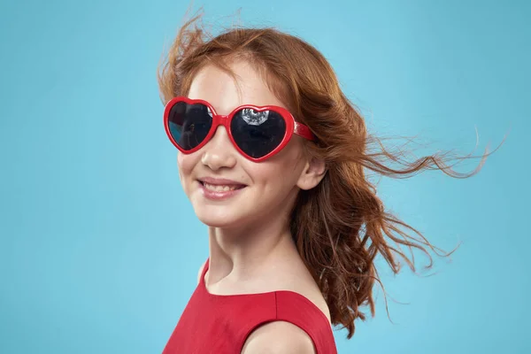 Bambina in occhiali da sole sotto forma di cuori vestito rosso capelli ricci sfondo blu — Foto Stock