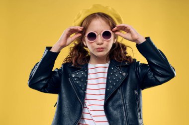 Güneş gözlüklü ve şapka takan neşeli küçük kız stüdyo sarı arka plan modası