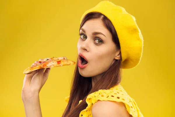 Mujer en sombrero amarillo con un pedazo de pizza en las manos sobre un fondo amarillo recortado vista de modelo de emoción — Foto de Stock