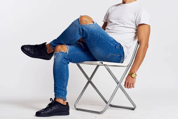 Un uomo in jeans e una t-shirt si siede su una sedia su uno sfondo leggero vista laterale — Foto Stock