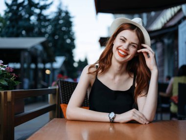 Beyaz şapkalı ve siyah elbiseli bir kadın caddedeki bir kafede oturuyor.