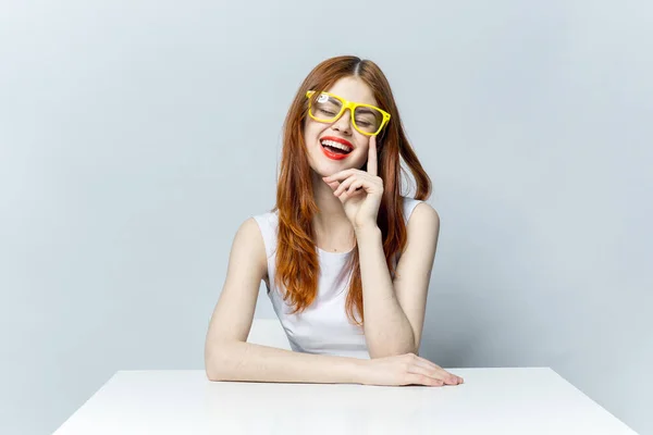 Mujer pelirroja sentada en una mesa con gafas amarillas sonrisa labios rojos modelo — Foto de Stock