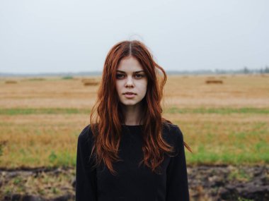 Siyah elbiseli, kızıl saçlı ve arka planda kuru bir tarla olan güzel bir kadın.