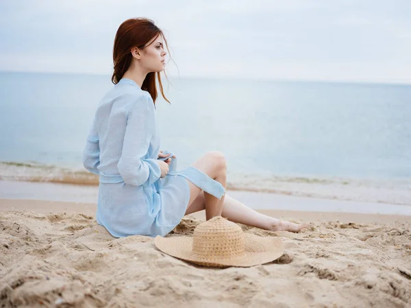 A woman in a blue dress on the sand and a hat in the hand of the ocean sea — Stock Photo, Image