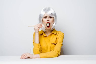 Beyaz peruklu güzel bir kadın sarı gömlekli bir masada oturur suşi sarmaları yer.