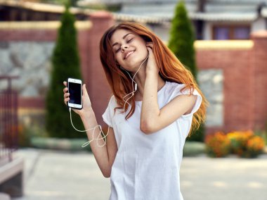 Beyaz tişörtlü neşeli bir kadın sokak müziğinde kulaklık takıyor.