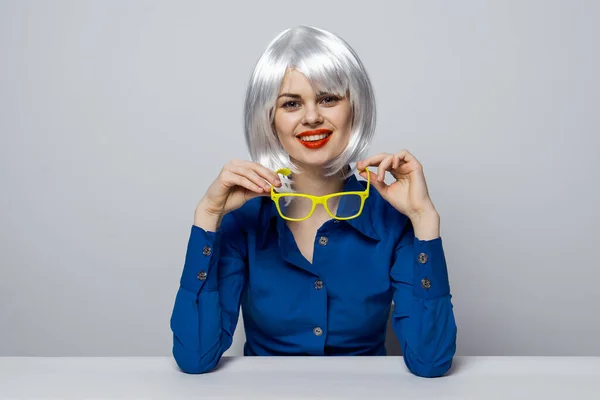 Mujer glamorosa sentada en una mesa en una camisa azul gafas amarillas en las manos encanto cosméticos — Foto de Stock