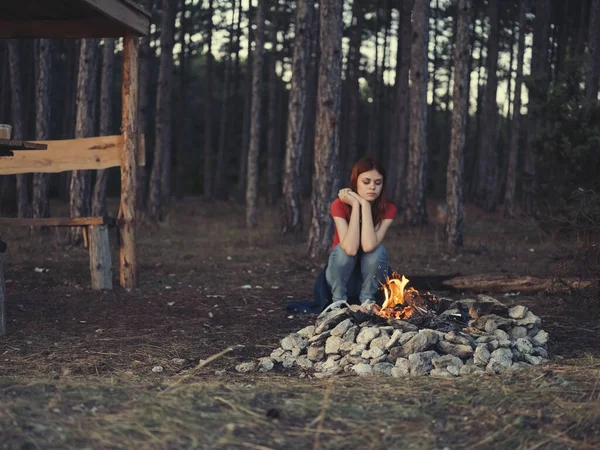 Una mujer romántica en jeans y una camiseta se sienta cerca de una fogata en la naturaleza —  Fotos de Stock