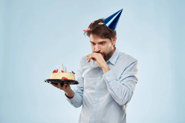 Hombre barbudo con un plato de pastel sobre un fondo azul y un sombrero de fiesta de cumpleaños en su cabeza — Foto de Stock