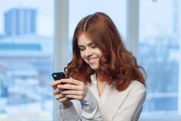 Mujer pelirroja en camisa con teléfono en las manos oficina tecnología oficial —  Fotos de Stock