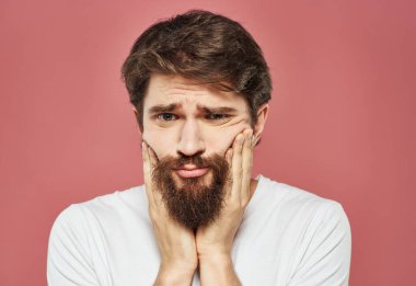 Sakallı bir adam pembe arka planda eli ile yüzüne dokunur.