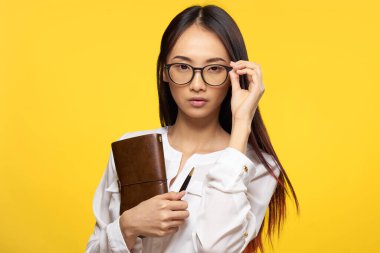 Gözlüklü bir iş kadını elinde not defteri ile resmi sarı izole bir arka plan tutuyor.