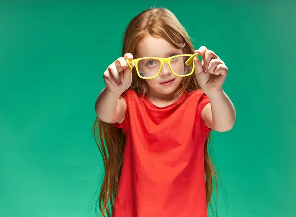 Dziewczyna z czerwonymi włosami trzymając żółte okulary w jej rękach zielone tło studio czerwony t-shirt — Zdjęcie stockowe