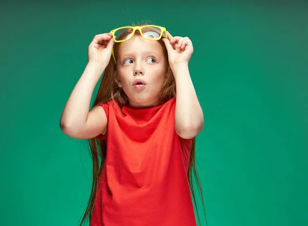 Meglepett lány piros haj tartó szemüveg a kezében érzelmek zöld háttér iskola — Stock Fotó