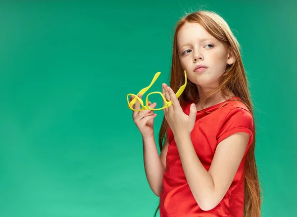 Κορίτσι με κόκκινα μαλλιά κρατώντας κίτρινα γυαλιά στα χέρια της πράσινο φόντο στούντιο κόκκινο t-shirt — Φωτογραφία Αρχείου