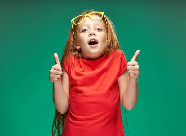 Alegre pelirroja chica sosteniendo gafas en sus manos aprendizaje escuela verde fondo —  Fotos de Stock