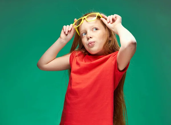 Vidám vörös hajú lány kezében szemüveg tanulás iskola zöld háttér — Stock Fotó