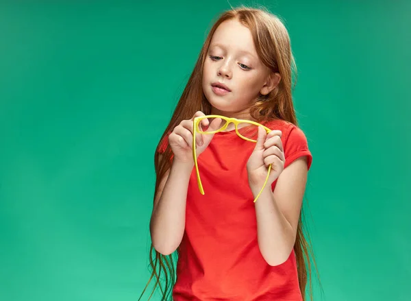 Chica con pelo rojo sosteniendo gafas amarillas en sus manos fondo verde estudio rojo camiseta — Foto de Stock