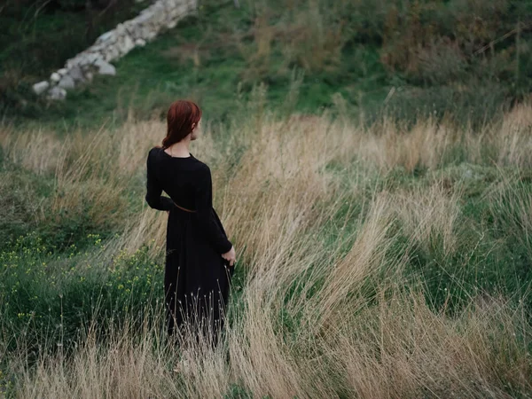 You are a woman in a black dress walking on dry grass in a meadow in the mountains to nature