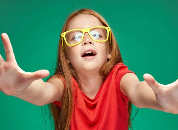 Χαριτωμένο κορίτσι με κόκκινα μαλλιά σε κίτρινα γυαλιά χειρονομίες με τα χέρια της συναισθήματα παιδική ηλικία Πράσινο φόντο — Φωτογραφία Αρχείου