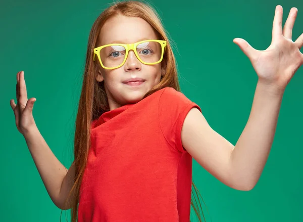 Mignonne fille aux cheveux roux en lunettes jaunes gestes avec ses mains émotions enfance Fond vert — Photo