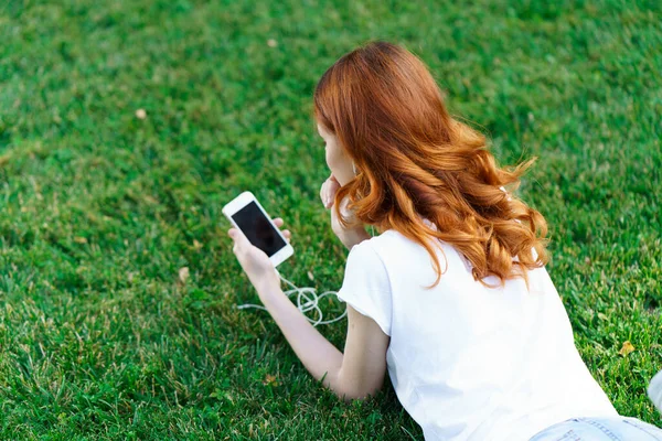 Kvinna ligger på gräsmattan med en telefon i händerna avkopplande i parken — Stockfoto