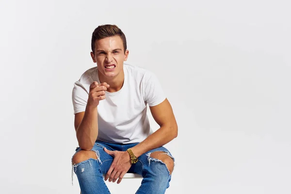 Man on a chair indoors torn jeans white t-shirt handsome face model light background — Stock Photo, Image