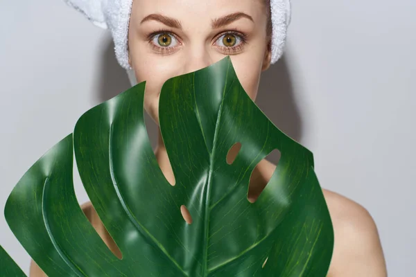 Una mujer con una toalla en la cabeza sostiene una hoja de palma cerca del maquillaje de la cara hombros desnudos cosmetología aspecto natural —  Fotos de Stock