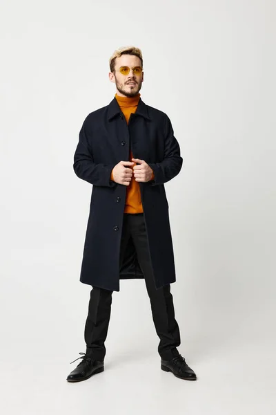 Fashionable man in a dark coat spread his legs shoulder-width apart on a light background and an orange sweater model — Stock Photo, Image