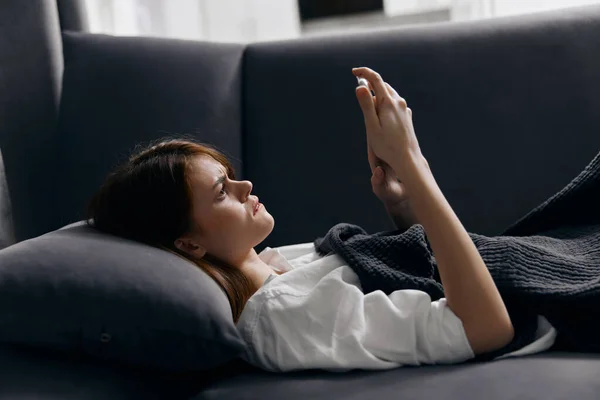 Kobieta pod kołdrą na sofie i telefon komórkowy w ręku — Zdjęcie stockowe