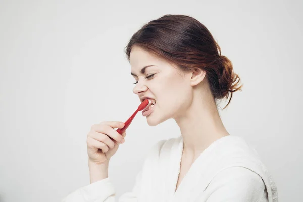 Woman with a toothbrush in her mouth on a light background and a white robe — Stock Photo, Image