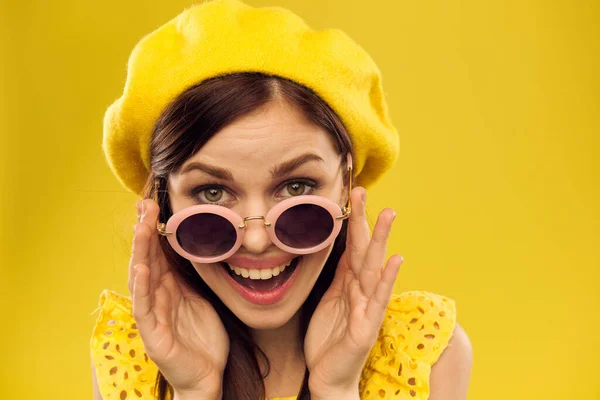 Sarı şapkalı ve şık gözlüklü mutlu bir kadın, sarı arka planda, duygularını eğlenceli bir şekilde yüzüne yakın tutuyor. — Stok fotoğraf