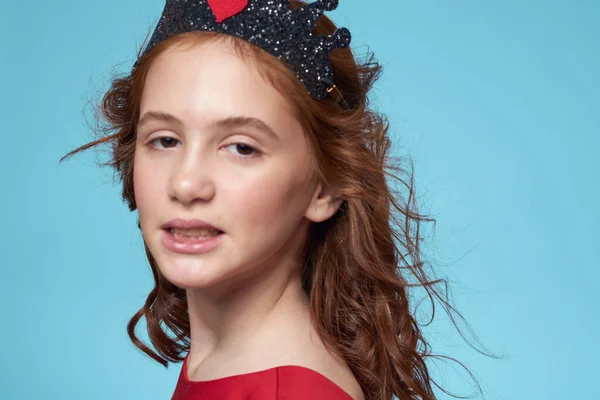 Bambina con i capelli ricci con una corona sulla testa vestito rosso Sorriso sfondo blu — Foto Stock