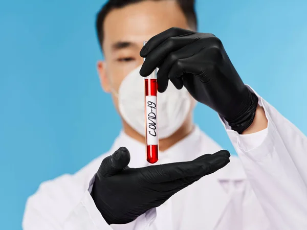 Un hombre de apariencia asiática en una máscara protectora prueba de sangre guantes negros diagnósticos fondo azul —  Fotos de Stock
