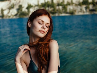 Kızıl saçlı kadın şeffaf bir nehrin kenarında ve arkasında dağlar var.