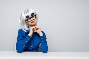 Beyaz peruklu, kırmızı dudaklı, siyah gözlüklü bir kadın masada oturuyor. 