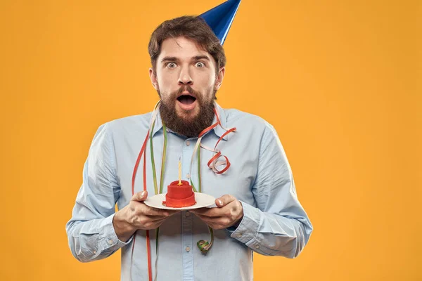 Wesoły mężczyzna z tortem na żółtym tle urodzinowa czapka na głowie — Zdjęcie stockowe