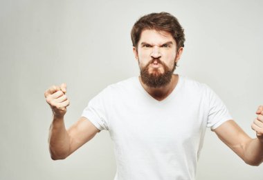 Beyaz tişörtlü öfkeli bir adam ellerini arka planda açık bir şekilde hareket ettiriyor.