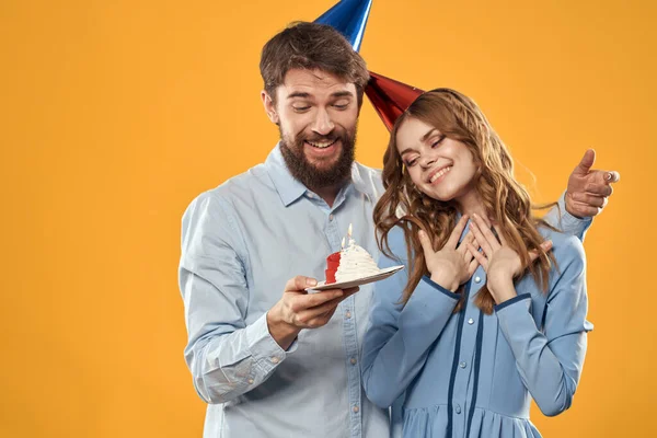 Birthday party man and woman on a yellow background in hats with a cake in their hands — Stock Photo, Image