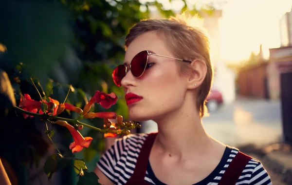 Vacker kvinna i glasögon nära röda blommor på gatan och röd solklänning randig t-shirt — Stockfoto