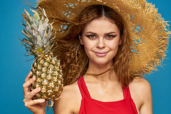 Uma mulher em um chapéu de palha com um abacaxi em suas mãos ou é um fundo de frutas exóticas azul divertido — Fotografia de Stock