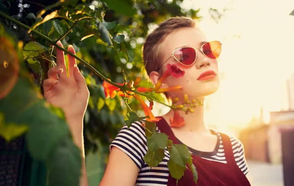 Belle femme dans des lunettes près de fleurs rouges sur la rue et rouge sundress rayé t-shirt — Photo