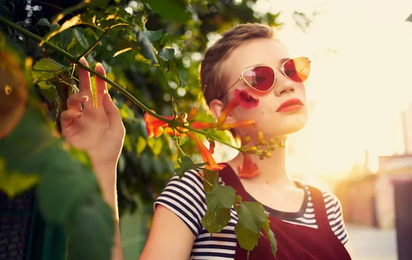 Vacker kvinna i glasögon nära röda blommor på gatan och röd solklänning randig t-shirt — Stockfoto