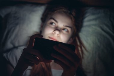 İnternette gece bağımlılığı sırasında elinde telefon olan bir kadın yatakta yatıyor.