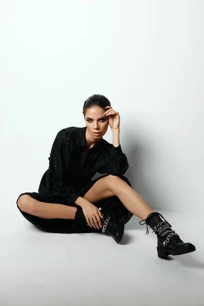 Mujer sentada en el suelo en un vestido negro botas de estudio de moda — Foto de Stock