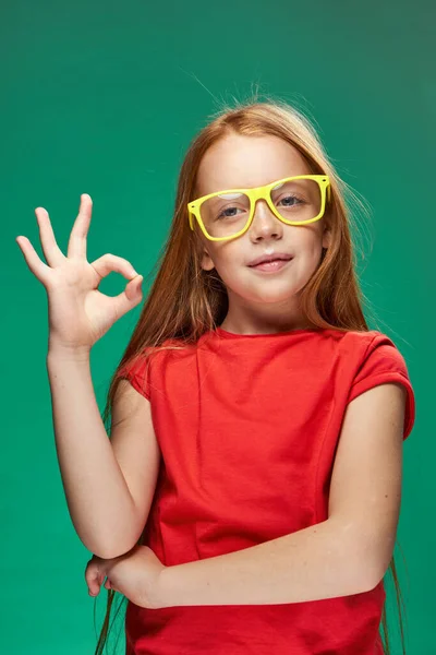 Κοκκινομάλλα κορίτσι με κίτρινα γυαλιά gesturing με τα χέρια της παιδική ηλικία μάθηση Πράσινο φόντο — Φωτογραφία Αρχείου